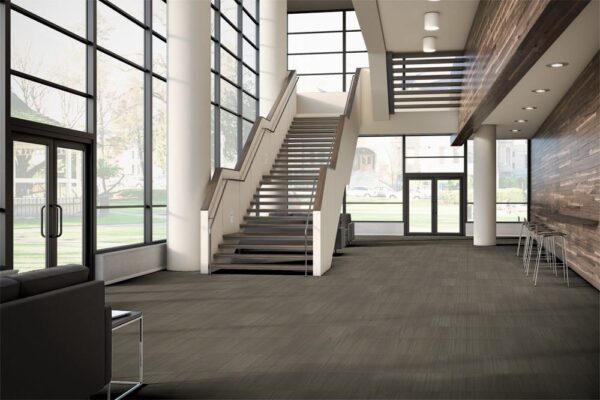 Non-fiction model flooring on the floor of a company lobby. A long set of stairs leads upward. Tall backed chairs are placed along the wall, opposite the door.