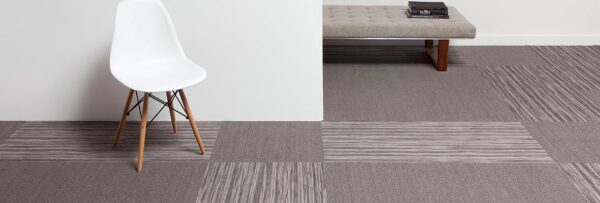Studio photography, looking into a room lined with solid colored carpeting, with Timber planks breaking up the design. A white plastic chair has been placed against the wall, with a long cushioned bench against the wall behind.