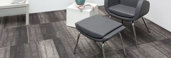 Studio shot of Well Versed model carpeting. The corner of this room contains a plush upholstered chair with matching ottoman. At the side is a white cube end table and a long table placed against the wall.