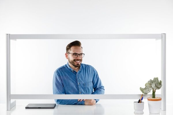 A man stands behind a counter, smiling, with a closed tablet next to him. A Counter Shield Wrap model screen separates him from the space in front.